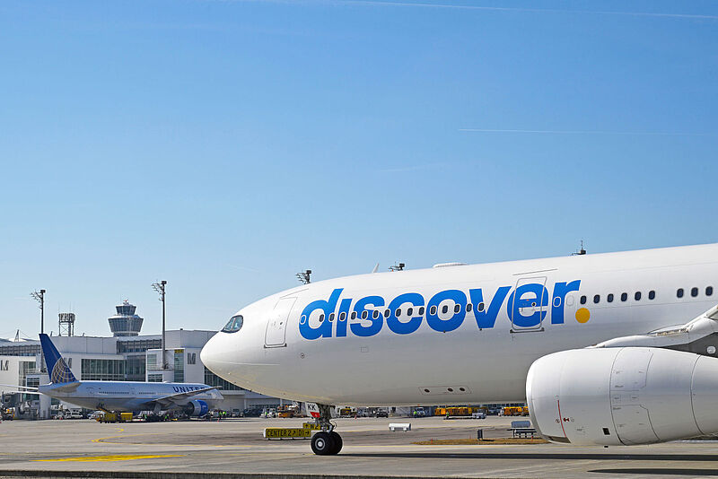  Ein Airbus-Flugzeug von Discover Airlines steht auf dem Vorfeld des Münchner Flughafens bei sonnigem Wetter mit blauen Himmel. Im Hintergrund sind Terminalgebäude und der Tower zu sehen.