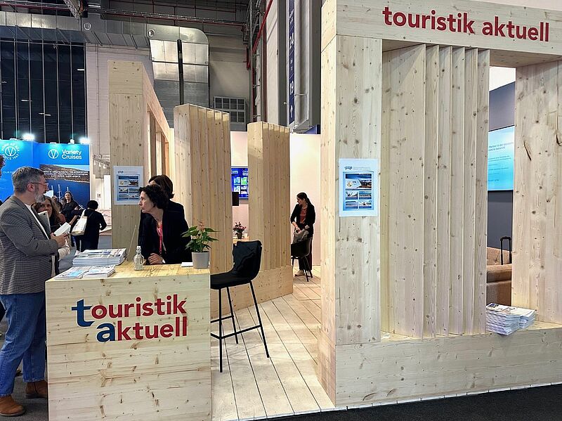 touristik aktuell ist traditionell in der Halle 25, Standnummer 127 Ein Messestand ganz aus Holz an dem drei Personen sind