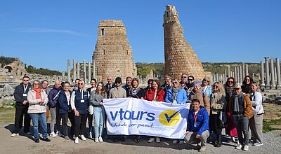 Bei strahlendem Sonnenschein präsentieren sich 20 Personen in der türkischen Ausgrabungsstätte Perge, sie halten eine Fahne des Reiseveranstalters Vtours