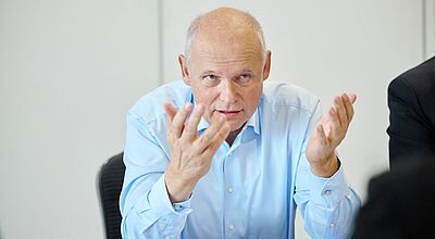 Ein Mann in hellblauem Hemd sitzt an einem Tisch, spricht gestikulierend mit erhobenen Händen und hat vor sich einige Dokumente liegen. Der Hintergrund ist schlicht und hell.