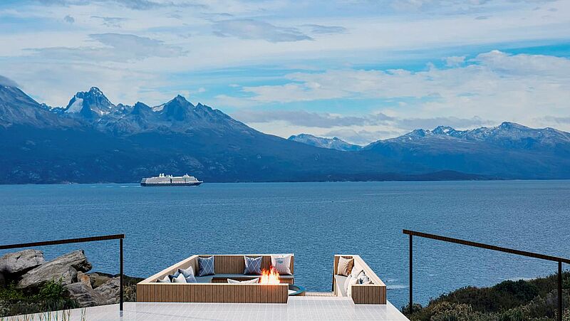 Das Anantara Ushuaia Patagonia Resort entsteht am Beagle-Kanal im Süden Argentiniens. Luxuriöse Terrasse mit Feuerstelle und Panoramablick auf ein Kreuzfahrtschiff im Beagle-Kanal vor schneebedeckten Anden in Patagonien – exklusives Reiseziel für Natur- und Kreuzfahrtliebhaber in Südamerika