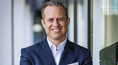 Ein Manager mittleren Alters in dunklem Jackett und weißem Hemd steht mit verschränkten Armen vor einem unscharfen Hintergrund. Der Hintergrund zeigt eine Fensterfront sowie Säulen einer Halle eines Bürogebäudes.