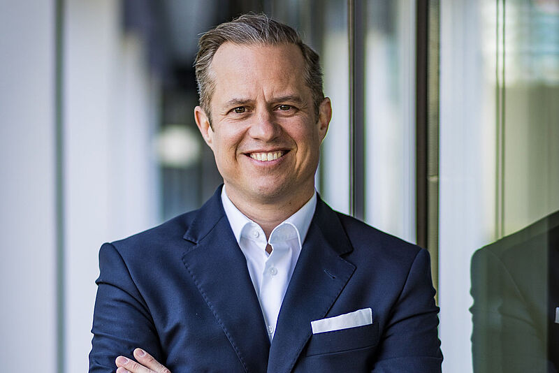 Bastian Gerlach wird bei Europcar Leiter des Key und Regional Account Managements Ein Manager mittleren Alters in dunklem Jackett und weißem Hemd steht mit verschränkten Armen vor einem unscharfen Hintergrund. Der Hintergrund zeigt eine Fensterfront sowie Säulen einer Halle eines Bürogebäudes.