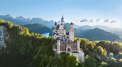 Luftaufnahme von Schloss Neuschwanstein, einem weißen Märchenschloss mit Türmen und Zinnen, das auf einem bewaldeten Hügel vor einer dramatischen Alpenkulisse thront. Im Hintergrund sind ein See und hohe Berge unter blauem Himmel zu sehen.