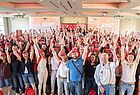 Eine große Gruppe von Menschen in einem Saal hebt die Hände in die Luft, viele tragen eine rote Baseball Cap mit dem Logo der Fluggesellschaft Emirates. Im Hintergrund ist auf zwei Leinwänden der Slogan „3… 2… Mainz!“ zu lesen. 