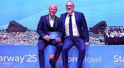 Zwei Männer sitzen auf einer Bühne und lächeln in die Kamera.