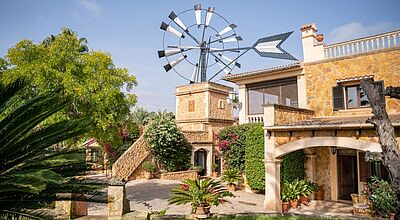 Mediterranes Landhaus auf Mallorca mit traditioneller Natursteinfassade, grünem Innenhof, Palmen, blühenden Pflanzen und historischem Windrad vor blauem Himmel – Ferienhaus oder Finca in ländlicher Idylle bei Palma.