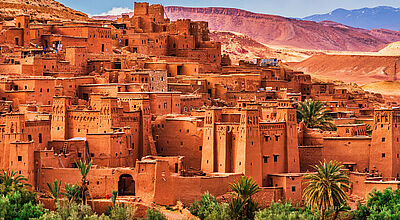 Gebäude einer traditionellen, marokkanischen Stadt in Lehmbauweise mit Palmen vor dem Hintergrund des Atlasgebirges