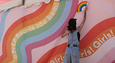 Buntes Regenbogen-Wandbild mit „Girls! Girls! Girls!“-Schriftzug. Zwei Personen posieren davor mit Regenbogenfächern.