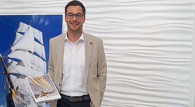 Ein lächelnder Reederei-Manager in den Mittvierzigern mit braunem Haar und Brille steht vor einem Roll-up mit einem weißen Segelkreuzfahrtschiff. Er trägt ein beiges Sakko, ein weißes Hemd und Jeand und hält in der Hand einen Kreuzfahrtkatalog.