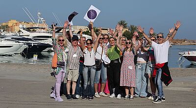 Ausgelassene Stimmung: Einer der Ausflüge führte nach El Gouna Eine fröhliche Gruppe von etwa fünfzehn Erwachsenen steht an einem sonnigen Yachthafen. Sie posieren eng beisammen, lachen und heben beide Arme jubelnd in die Luft. Im Hintergrund liegen mehrere weiße Motorboote im Wasser, dahinter Palmen und Gebäude. Alle tragen farbenfrohe oder sommerliche Kleidung sowie Namensschilder, einige halten Papier oder Wasserflaschen in der Hand