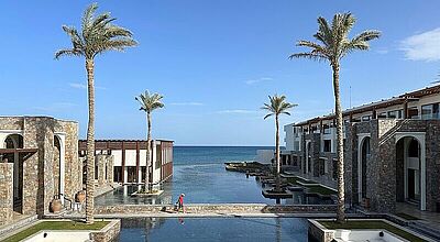 Blick über einen Pool auf das Meer, rechts und links Hotelgebäude