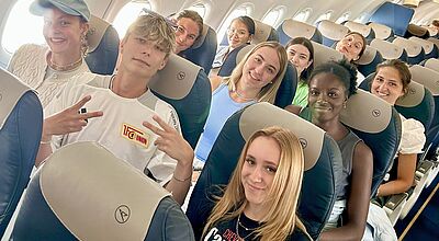 Eine Gruppe junger Menschen sitzt lächelnd in den Sitzen eines Flugzeugs. Sie wirken fröhlich und entspannt, einige zeigen das Peace-Zeichen. Die Kabine ist hell und freundlich beleuchtet, im Hintergrund sind weitere Fluggäste zu sehen.
