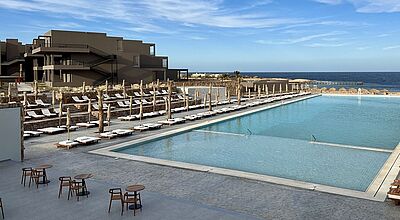 Luxuriöse Poolanlage eines Resorts mit zahlreichen weißen Sonnenliegen und geschlossenen Sonnenschirmen entlang des Wassers. Im Hintergrund befindet sich ein modernes, dunkel gehaltenes Gebäude mit Außentreppen sowie das blaue Meer mit einem Steg. Der Himmel ist klar und sonnig. Die Szenerie strahlt Ruhe und Exklusivität aus.