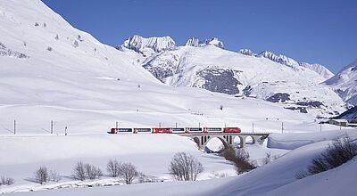 Ein roter Zug des Glacier Express fährt über ein steinernes Viadukt durch eine verschneite Berglandschaft in den Schweizer Alpen. Die Szenerie ist geprägt von tiefem Schnee, kahlen Bäumen und imposanten schneebedeckten Gipfeln unter klarem, blauem Himmel.