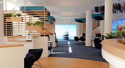Blick in den neuen, modernen TUI-Flagshipstore in Aschaffenburg Innenansicht des neuen TUI-Flagshipstores in Aschaffenburg: moderne Beratungslounge mit hellen Holzelementen, blauen Stoffdecken im Wellen-Design, Pflanzen und offenen Beratungsplätzen. Im Hintergrund ein großes Bildschirmbild mit einem Kreuzfahrtschiff.