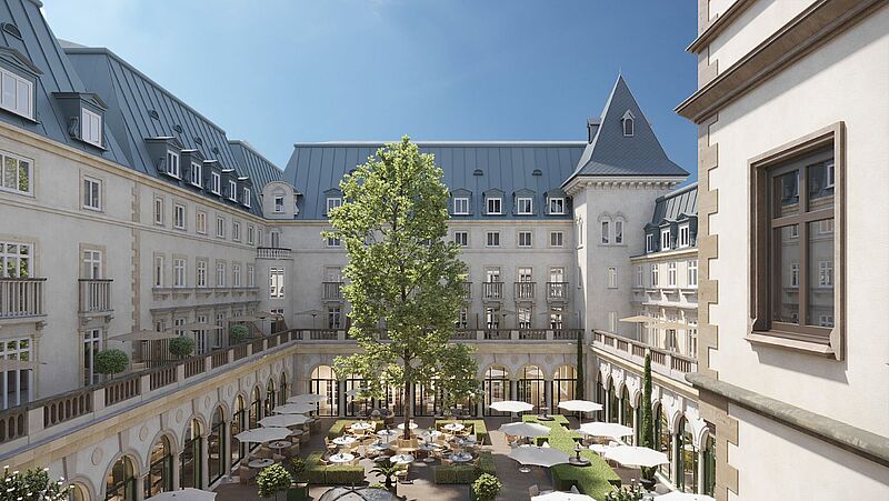 Innenhof eines historischen Gebäudes mit heller Fassade, Mansardendach und zentralem Baum, umgeben von Sitzbereichen und Sonnenschirmen unter blauem Himmel.