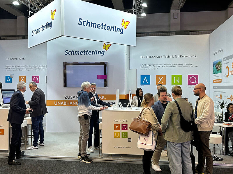 Messestand von Schmetterling auf einer Reisemesse. Mehrere Personen stehen und unterhalten sich vor weißen Wänden mit Firmenlogo und bunten Symbolen für Softwarelösungen (A, X, Q, U, N). Auf den Wänden sind Werbetexte zu Full-Service-Technik für Reisebüros zu sehen. Links arbeiten zwei Männer an Computern, rechts führen Besucher Gespräche.