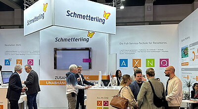 Messestand von Schmetterling auf einer Reisemesse. Mehrere Personen stehen und unterhalten sich vor weißen Wänden mit Firmenlogo und bunten Symbolen für Softwarelösungen (A, X, Q, U, N). Auf den Wänden sind Werbetexte zu Full-Service-Technik für Reisebüros zu sehen. Links arbeiten zwei Männer an Computern, rechts führen Besucher Gespräche.