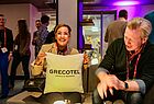 Ein Frau und ein Mann sitzen in einer Bar. Die Frau hält ein Kissen in der Hand mit der Aufschrift Grecotel.