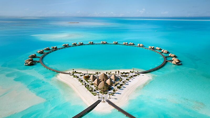 An einem kreisrunden Steg im türkisblauen Wasser befinden sich viele Overwater-Villen