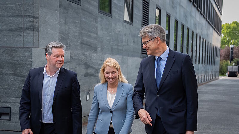 Ein Manager, ein Politiker und eine Managerin gehen zusammen in Business-Kleidung vor einem grauen Bürogebäude aus Beton mit Fensterfassade entlang.