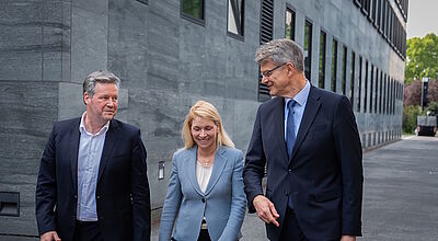Ein Manager, ein Politiker und eine Managerin gehen zusammen in Business-Kleidung vor einem grauen Bürogebäude aus Beton mit Fensterfassade entlang.
