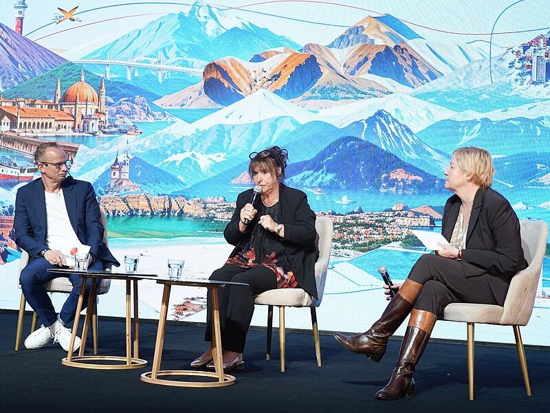 Auf der Bühne einer Tagung spricht eine Rednerin ins Mikro, eingerahmt von einem Manager und einer Pressesprecherin.