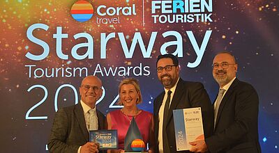 Drei Herren in Anzügen stehen auf einer Bühne und gratulieren einer Dame in einem pinken Kleid, die einen Preis entgegennimmt. 