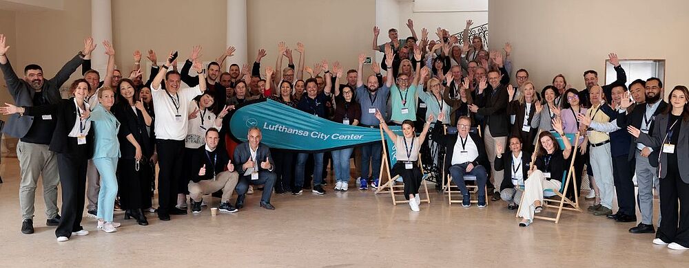 Eine große Gruppe von Teilnehmerinnen und Teilnehmern der Lufthansa City Center Vollversammlung posiert in einem hellen Konferenzraum. Alle lächeln, heben die Arme zum Gruß und stehen oder sitzen in mehreren Reihen. In der Mitte halten zwei Personen ein türkisfarbenes Banner mit der Aufschrift „Lufthansa City Center“.