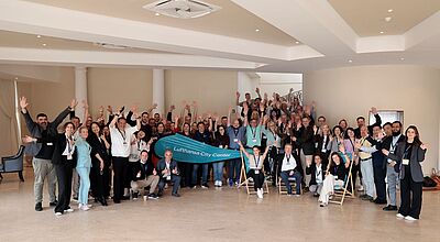 Eine große Gruppe von Teilnehmerinnen und Teilnehmern der Lufthansa City Center Vollversammlung posiert in einem hellen Konferenzraum. Alle lächeln, heben die Arme zum Gruß und stehen oder sitzen in mehreren Reihen. In der Mitte halten zwei Personen ein türkisfarbenes Banner mit der Aufschrift „Lufthansa City Center“.