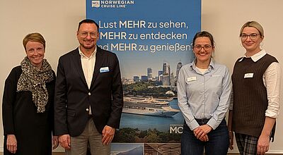 Vier Personen stehen lächelnd vor einem blauen Banner von Norwegian Cruise Line mit dem Schriftzug „Lust MEHR zu sehen, MEHR zu entdecken und MEHR zu genießen“. Die Gruppe besteht aus drei Frauen und einem Mann. Alle tragen Namensschilder und sind geschäftlich gekleidet. Das Banner zeigt im Hintergrund eine Skyline, ein Kreuzfahrtschiff und eine Küstenlandschaft.