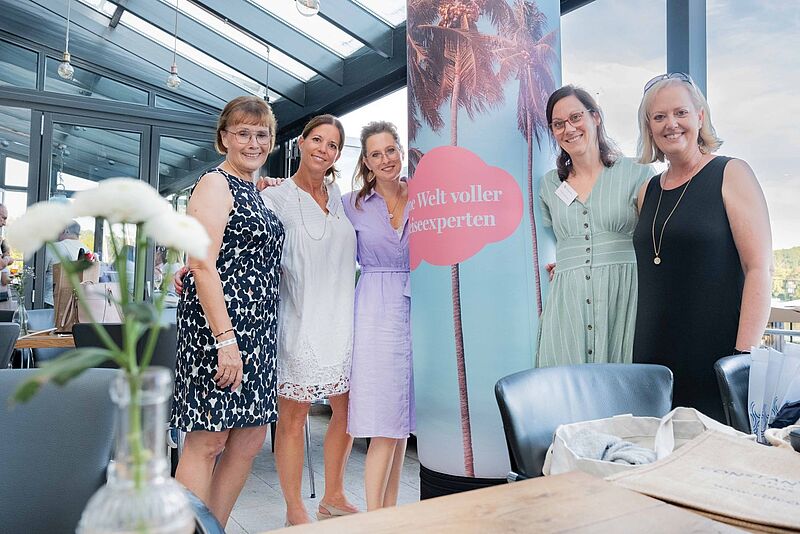Sechs Frauen stehen in einer hellen, modernen Location nebeneinander und posieren für ein Gruppenfoto. Im Vordergrund steht eine weiß blühende Blume in einer Vase, und im Hintergrund ist eine Säule mit einem Motiv aus Palmen und einer roten Wolke mit dem Schriftzug „Eine Welt voller Reiseexperten“ sichtbar. Die Atmosphäre wirkt freundlich und sommerlich.