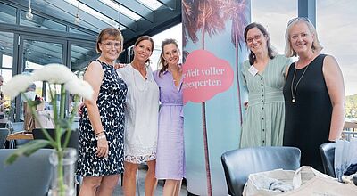 Sechs Frauen stehen in einer hellen, modernen Location nebeneinander und posieren für ein Gruppenfoto. Im Vordergrund steht eine weiß blühende Blume in einer Vase, und im Hintergrund ist eine Säule mit einem Motiv aus Palmen und einer roten Wolke mit dem Schriftzug „Eine Welt voller Reiseexperten“ sichtbar. Die Atmosphäre wirkt freundlich und sommerlich.