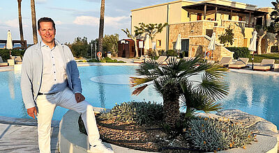 Ein lächelnder Mann, es ist Benjamin Jacobi von TUI Deutschland, steht am Rand eines eleganten Pools im Grecotel Imperial auf Kos. Er trägt ein hellblaues Sakko, ein helles Hemd, weiße Hose und weiße Sneaker. Eine seiner Füße ruht auf dem erhöhten, runden Pflanzbeet mit einer kleinen Palme und Bodendeckern. Im Hintergrund sind Sonnenliegen, Sonnenschirme, Palmen und ein modernes Gebäude mit Natursteinfassade und Terrasse zu sehen. Die Sonne wirft lange Schatten, was auf den späten Nachmittag hinweist.