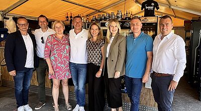 Eine Gruppe von acht Personen steht lächelnd nebeneinander in einem Zelt mit Bar im Hintergrund. Die Personen sind sommerlich bis geschäftlich leger gekleidet. Im Hintergrund stehen Getränke und Keramikkrüge auf Regalen, über dem Tresen hängt ein Trikot von Eintracht Frankfurt mit der Aufschrift „Indeed“. Die Stimmung wirkt freundlich und ungezwungen.