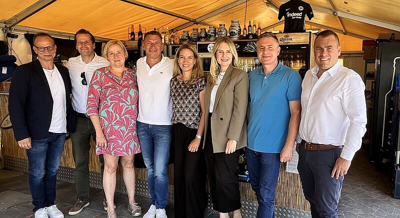 Diskutierten über die Zukunft von B4C (von links): Cornelius Meyer, Markus Zahn, Beate Zwermann, Steven Mathes, Anja Dammann, Katharina Port, Deniz Ugur und Florian Hartmann. Foto: uf Eine Gruppe von acht Personen steht lächelnd nebeneinander in einem Zelt mit Bar im Hintergrund. Die Personen sind sommerlich bis geschäftlich leger gekleidet. Im Hintergrund stehen Getränke und Keramikkrüge auf Regalen, über dem Tresen hängt ein Trikot von Eintracht Frankfurt mit der Aufschrift „Indeed“. Die Stimmung wirkt freundlich und ungezwungen.