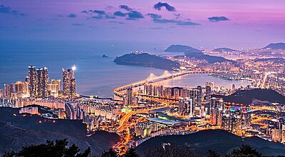 Blick von einem Berg auf die Millionenmetropole Busan im Abendlicht. Die Sonne ist bereits untergegangen, der Himmel aber noch dunkelviolett. 