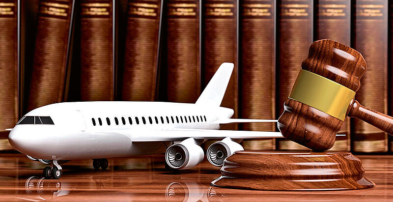 Ein Flugzeugmodell steht zusammen mit einem Richterhammer auf einem Holztisch, im Hintergrund sind Bücher zu sehen.