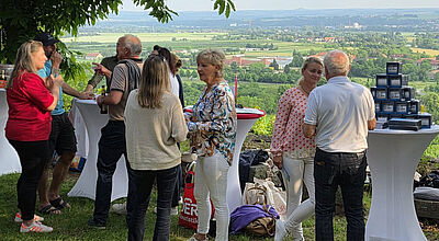 Eine Gruppe von Menschen steht im Freien an Stehtischen und unterhält sich bei einem Treffen oder Event. Im Hintergrund ist eine weite Landschaft mit Feldern, Bäumen und Gebäuden zu sehen. Auf einem der Tische sind gestapelte kleine Kartons und Prospekte mit einem Schild des Reiseveranstalters LMX. Einige Taschen am Boden tragen Logos von DERTOUR GROUP und Eberhardt Travel, was auf eine Veranstaltung mit touristischen Partnern hinweist.