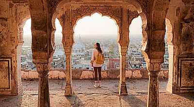Eine Person mit Rucksack steht in einem alten, kunstvoll verzierten Torbogen und blickt über eine Stadtlandschaft mit historischen Gebäuden bei warmem Abendlicht.