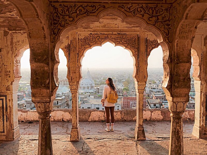 Eine Person mit Rucksack steht in einem alten, kunstvoll verzierten Torbogen und blickt über eine Stadtlandschaft mit historischen Gebäuden bei warmem Abendlicht.