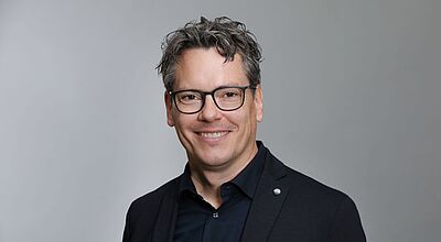 Ein Mann mit schwarzer Brille und schwarzem Hemd lächelt in die Kamera