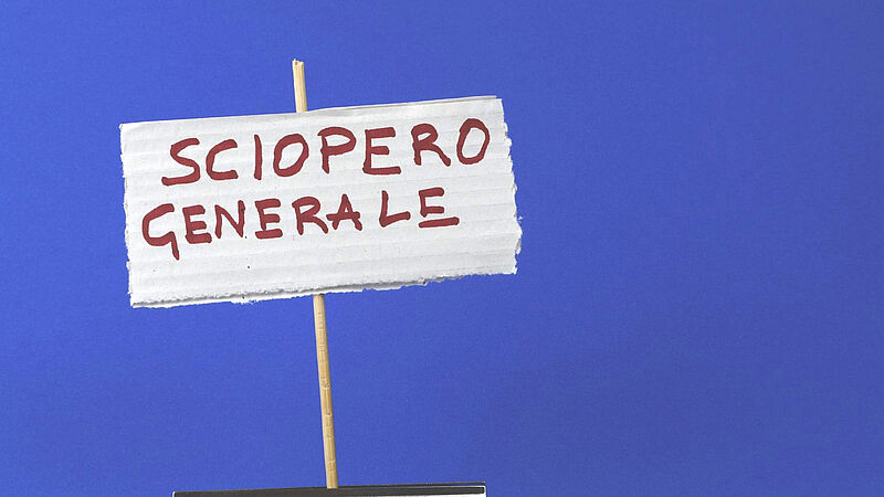 Handgemachtes Schild aus Wellpappe mit der roten Aufschrift „Sciopero Generale“ (italienisch für „Generalstreik“), befestigt an einem dünnen Holzstab, vor blauem Hintergrund.