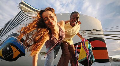 Die Kussmundflotte feiert 2026 ihr 30-jähriges Bestehen. Foto: Max Threlfall An der Pier vor dem Bug eines großen Kreuzfahrtschiffes, auf dem der charakteristische rote Kussmund der Reederei Aida zu sehen ist, beugt sich ein junges Paar in die Kamera. Sie halten sich an der Hand, die Frau hat lange gelockte braune Haare und ein Top an, der Mann trägt kurze schwarze Haare und in der Hand einen Rucksack. Beide strahlen.