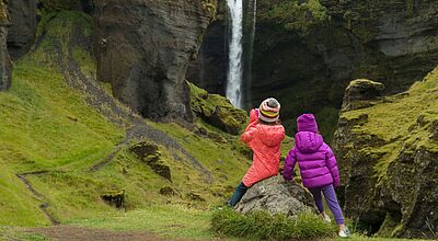 Zwei Mädchen in Rückansicht mit Mützen und Winterjacken schauen auf einen Wasserfall, der von grün bewachsenen Felsen herunterstürzt.