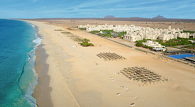 Am langen Sandstrand im Süden der Insel Boa Vista liegt das Riu Touareg, das erste Hotel von RIU auf der Insel Boa Vista Luftaufnahme eines langen, goldgelben Sandstrandes mit türkisblauem Meer auf der linken Seite. Am breiten Strand sind Sonnenliegen und Strohdach-Sonnenschirme aufgestellt. Im Hintergrund liegt ein großes, helles Hotelresort mit mehreren Gebäuden, umgeben von Wüste und flachem Gebirgszug unter blauem Himmel.