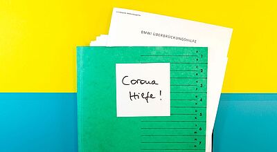 Im Dezember finden gleich zwei Webinare zum Thema Überbrückungshilfen statt Ein grüner Aktenordner liegt vor gelbem und blauem Hintergrund, darauf klebt ein Zettel mit der Aufschrift „Corona Hilfe“, und oben ragt ein Formular mit dem Titel „BMWi Überbrückungshilfe“ heraus – Symbolbild für staatliche Coronahilfen und Überbrückungshilfe während der Pandemie.