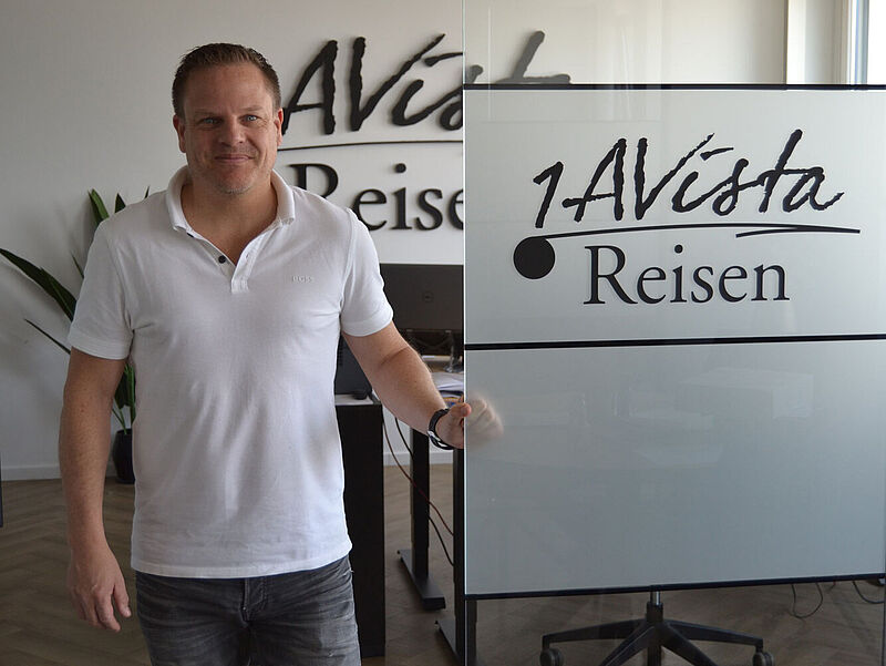 Ein Geschäftsführer mittleren Alters mit kurzen braunen Haaren, blauer Jeans und weißem Poloshirt steht in einer Firmenzentrale. Auf einer Bürotür aus Milchglas prangt der Firmenname 1A Vista Reisen.