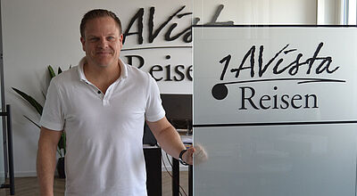 Ein Geschäftsführer mittleren Alters mit kurzen braunen Haaren, blauer Jeans und weißem Poloshirt steht in einer Firmenzentrale. Auf einer Bürotür aus Milchglas prangt der Firmenname 1A Vista Reisen.